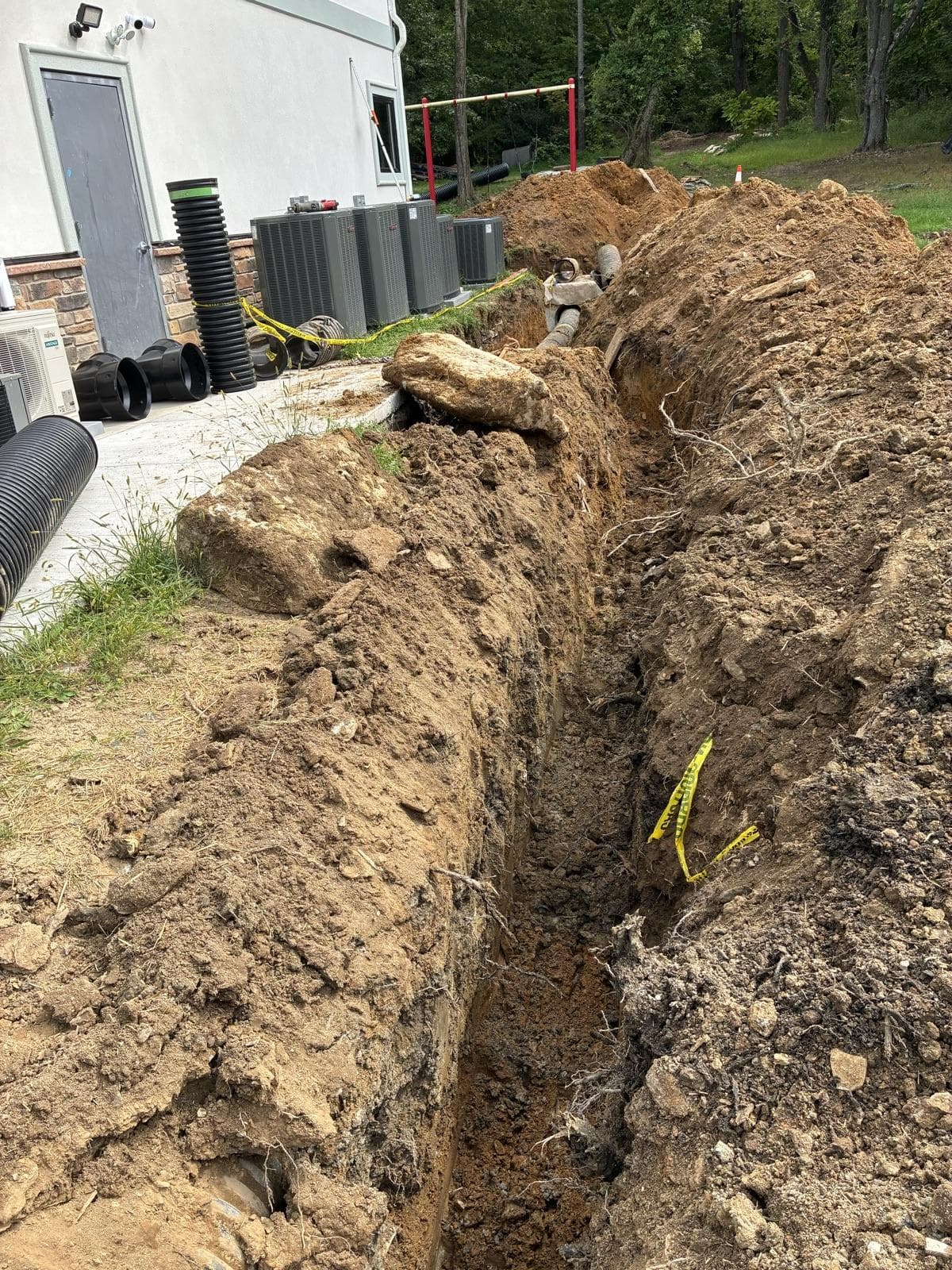 A long, straight trench dug along the side of a building, with drainage pipe staged beside it.