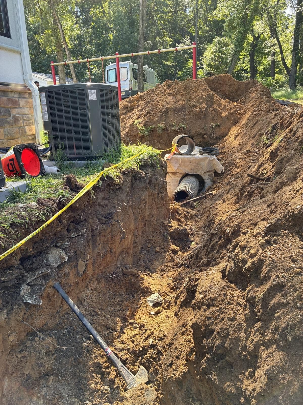 A deep trench dug right beside a house's air-conditioning unit, with a catch basin and pipe in the cut.